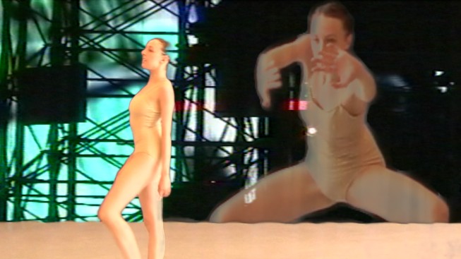 Dancer Dona Wiley in a nude leotard bevels her foot looking straight ahead. There is a projected image of her in a crouch behind her.